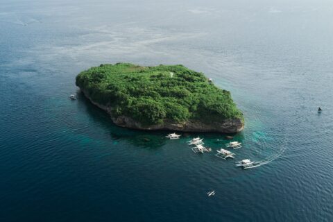 Vista aérea de la isla Pescador, una roca cubierta de vegetación verde con su faro blanco, rodeada de una decena de bancas de buceo sobre un mar azul oscuro