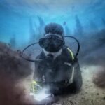 Un buceador con traje negro, tumbado sobre un fondo de arena entre dos rocas cubiertas de algas pardas, escribe en una pizarra blanca, con siluetas de laminarias recortadas en el azul detrás de él.