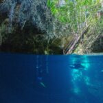 Vista mitad aire, mitad agua en la entrada del cenote The Pit: la bóveda de caliza y la escalera de madera bajo la selva arriba, y dos buceadores en el agua azul profundo abajo