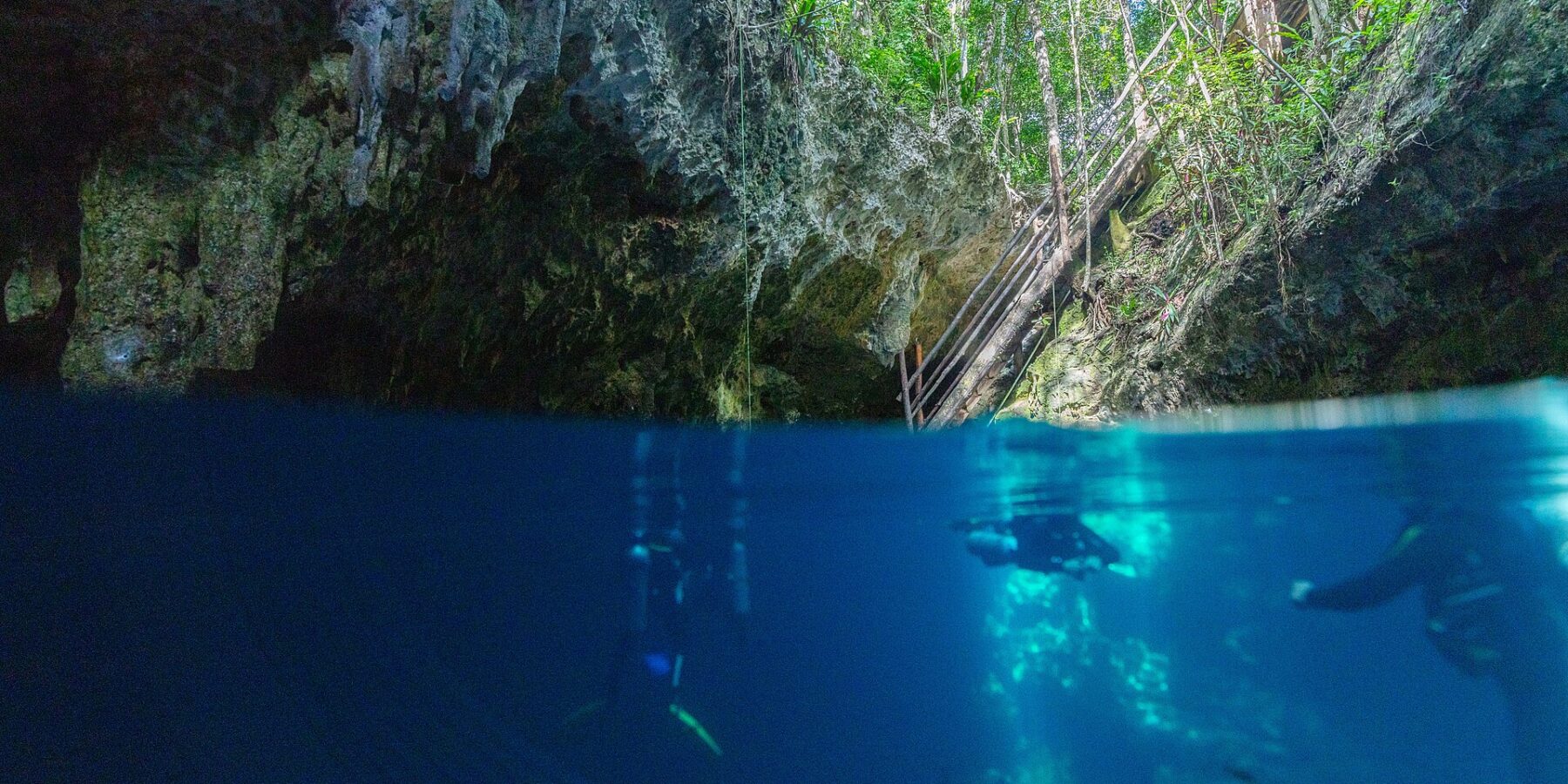 Vista mitad aire, mitad agua en la entrada del cenote The Pit: la bóveda de caliza y la escalera de madera bajo la selva arriba, y dos buceadores en el agua azul profundo abajo