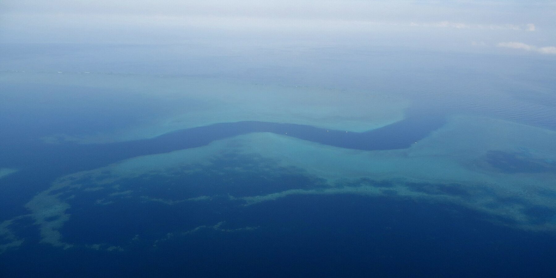 Vista aérea de un canal azul oscuro en forma de S que serpentea entre las plataformas turquesa de la barrera de coral, con algunos barcos blancos pequeños en el paso