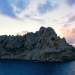 La isla Maïre, una mole de caliza clara con crestas dentadas, se perfila a contraluz sobre un cielo de atardecer con nubes rosas y malvas, encima de un mar azul oscuro.