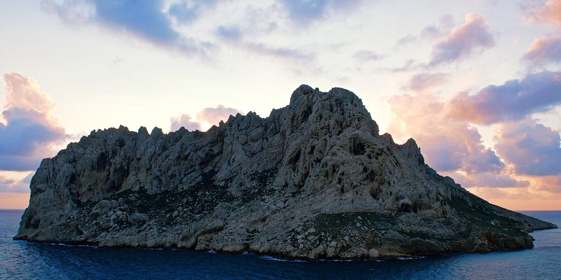La isla Maïre, una mole de caliza clara con crestas dentadas, se perfila a contraluz sobre un cielo de atardecer con nubes rosas y malvas, encima de un mar azul oscuro.