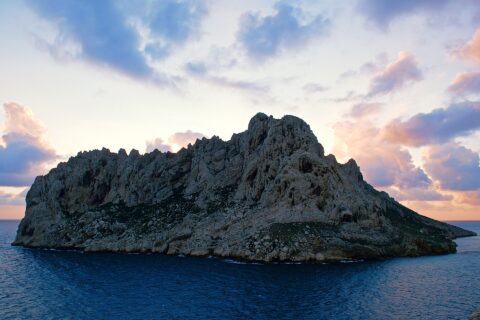 La isla Maïre, una mole de caliza clara con crestas dentadas, se perfila a contraluz sobre un cielo de atardecer con nubes rosas y malvas, encima de un mar azul oscuro.