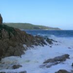 Las olas rompen en espuma blanca sobre las rocas de granito de L'Escalet, en primer plano, frente a un mar azul oscuro y agitado, con la costa boscosa del cabo Camarat y su faro blanco a lo lejos.