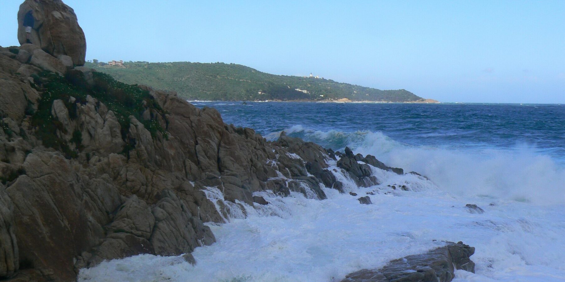 Las olas rompen en espuma blanca sobre las rocas de granito de L'Escalet, en primer plano, frente a un mar azul oscuro y agitado, con la costa boscosa del cabo Camarat y su faro blanco a lo lejos.