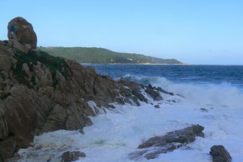 Las olas rompen en espuma blanca sobre las rocas de granito de L'Escalet, en primer plano, frente a un mar azul oscuro y agitado, con la costa boscosa del cabo Camarat y su faro blanco a lo lejos.
