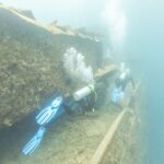 Dos buceadores recorren la cubierta del Thistlegorm a lo largo de un pasillo, en un agua de azul lechoso