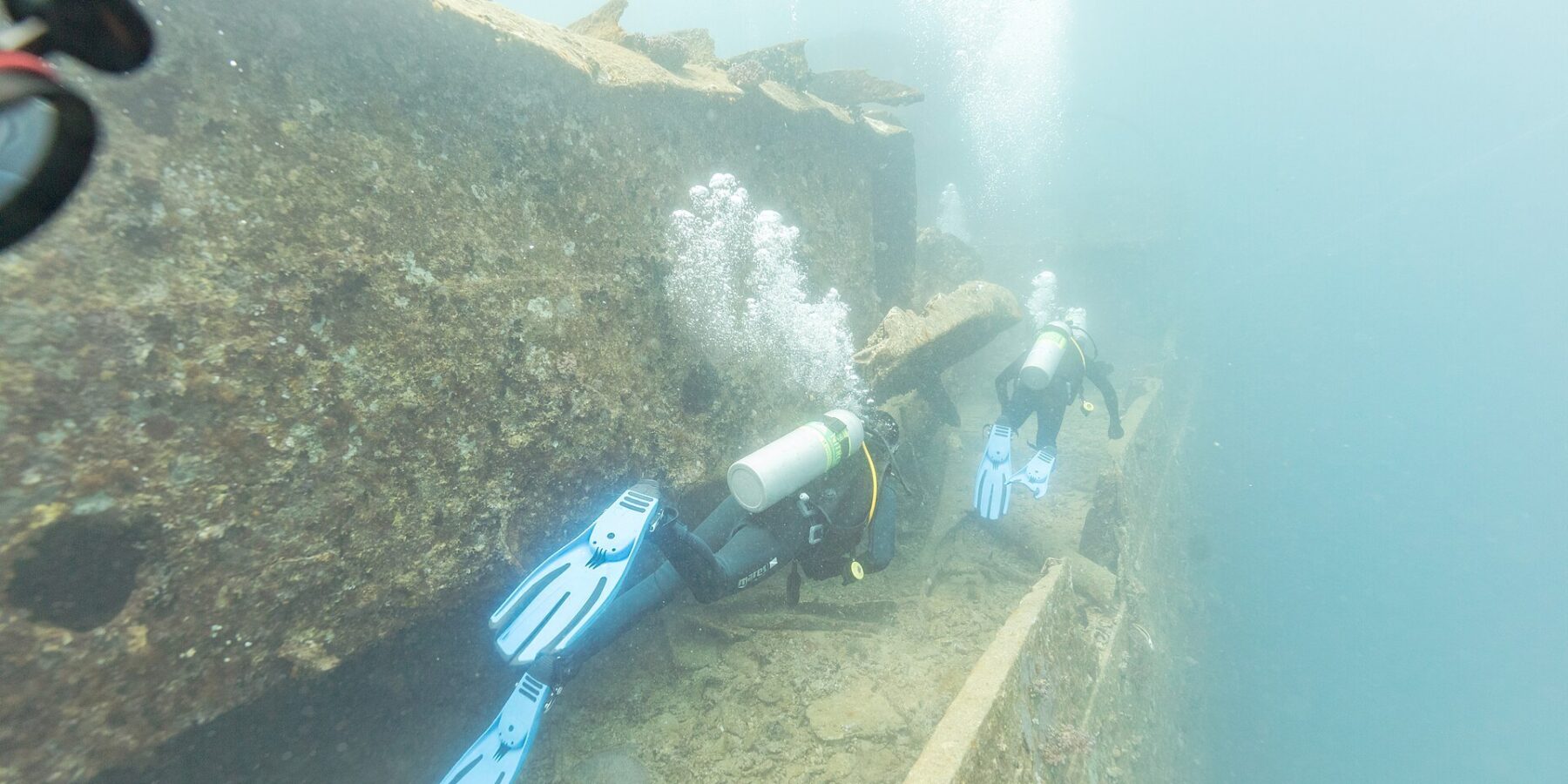 Dos buceadores recorren la cubierta del Thistlegorm a lo largo de un pasillo, en un agua de azul lechoso