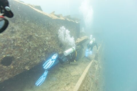 Dos buceadores recorren la cubierta del Thistlegorm a lo largo de un pasillo, en un agua de azul lechoso