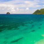 Vista aérea de la larga playa de Le Diamant bordeada de agua turquesa, con la Roca del Diamante mar adentro y el morne Larcher, una colina verde, a la derecha