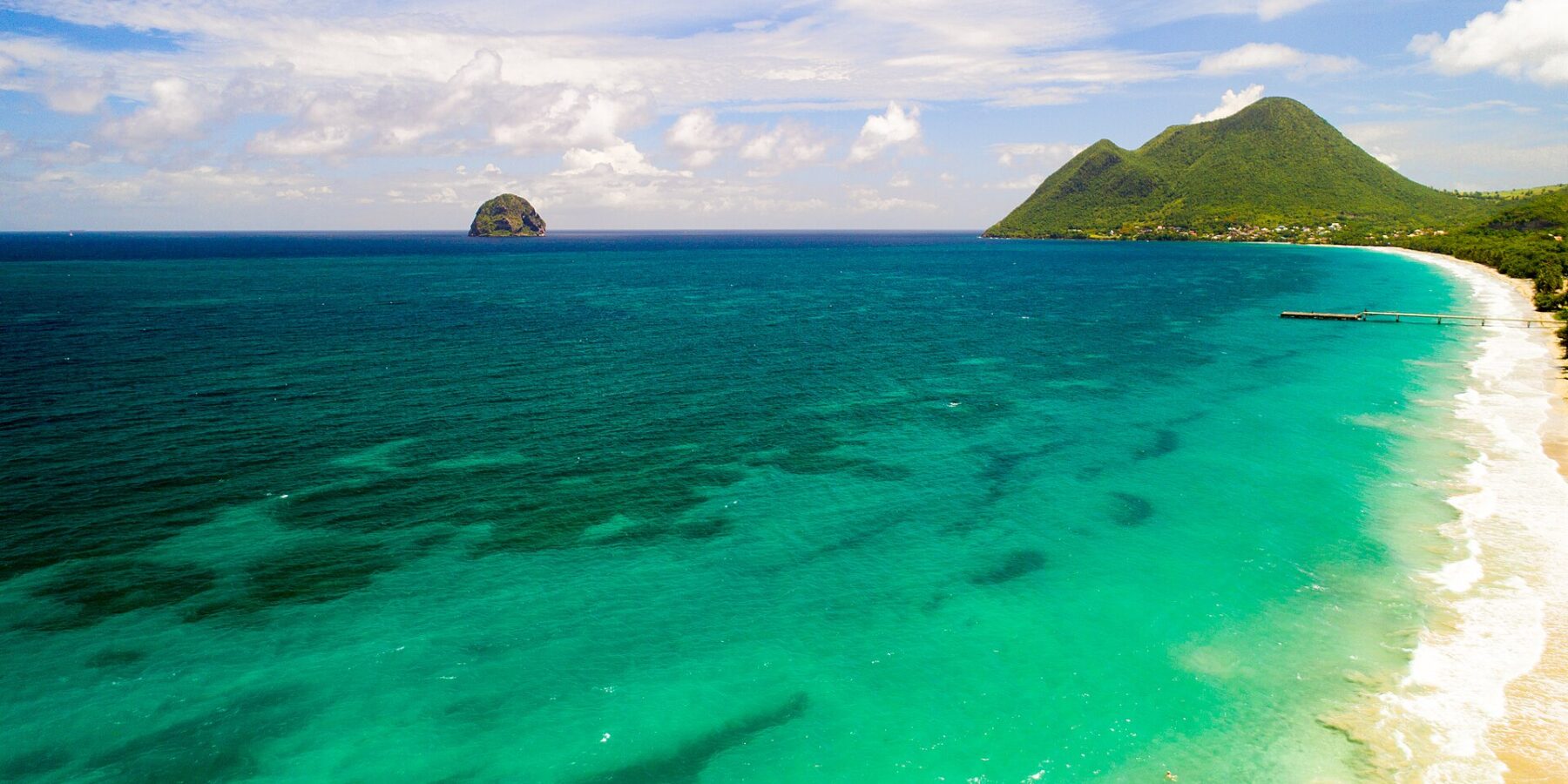 Vista aérea de la larga playa de Le Diamant bordeada de agua turquesa, con la Roca del Diamante mar adentro y el morne Larcher, una colina verde, a la derecha