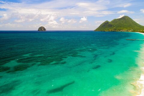 Vista aérea de la larga playa de Le Diamant bordeada de agua turquesa, con la Roca del Diamante mar adentro y el morne Larcher, una colina verde, a la derecha