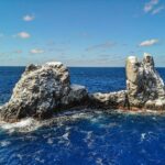 Roca Partida vista desde un dron: una roca blanqueada por las aves, partida en dos picos y batida por un mar azul oscuro bajo un cielo azul salpicado de nubes