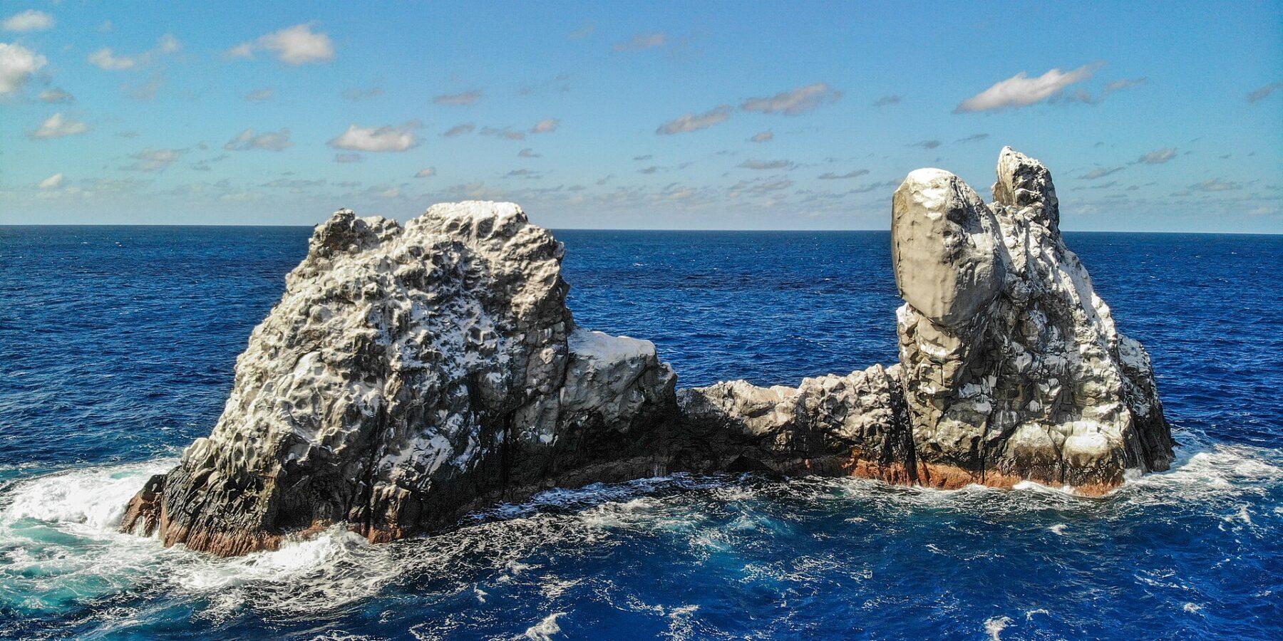 Roca Partida vista desde un dron: una roca blanqueada por las aves, partida en dos picos y batida por un mar azul oscuro bajo un cielo azul salpicado de nubes