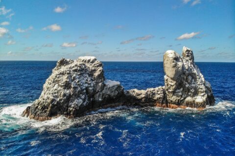 Roca Partida vista desde un dron: una roca blanqueada por las aves, partida en dos picos y batida por un mar azul oscuro bajo un cielo azul salpicado de nubes