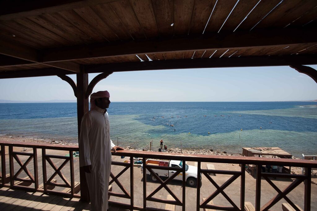 Desde una terraza de madera, un hombre con galabiya mira la poza del Blue Hole, rodeada de arrecife claro, donde nadan unos bañistas