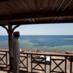 Desde una terraza de madera, un hombre con galabiya mira la poza del Blue Hole, rodeada de arrecife claro, donde nadan unos bañistas