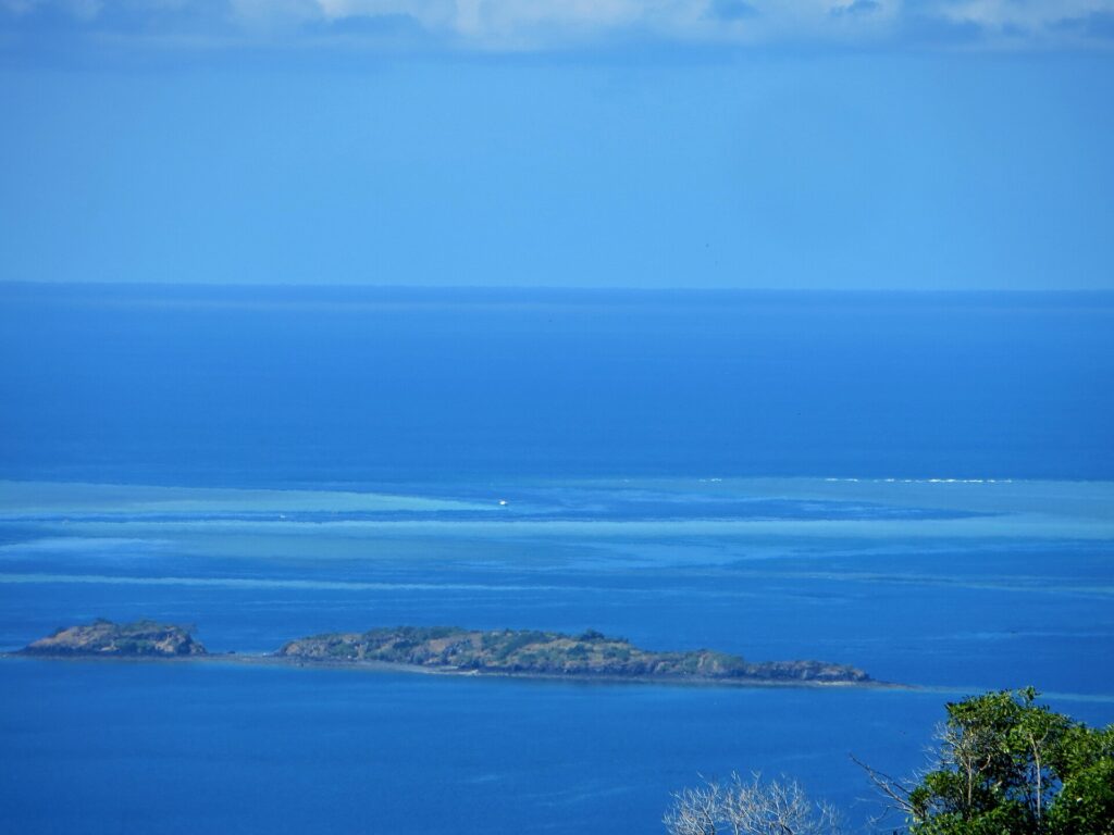 Vista desde lo alto de la laguna azul, con dos islotes rocosos en primer plano y, a lo lejos, las plataformas claras de la barrera de coral donde un barco blanco está parado en el paso