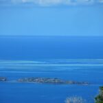 Vista desde lo alto de la laguna azul, con dos islotes rocosos en primer plano y, a lo lejos, las plataformas claras de la barrera de coral donde un barco blanco está parado en el paso