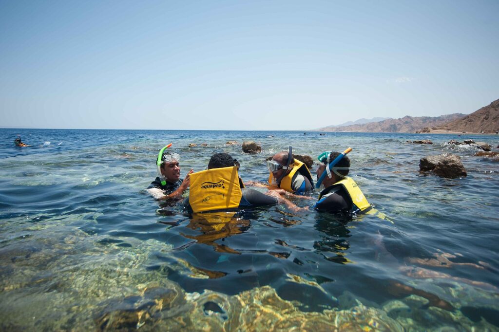 Nadadores con chalecos amarillos, máscaras y tubos en el agua clara del Blue Hole, con las montañas del Sinaí detrás